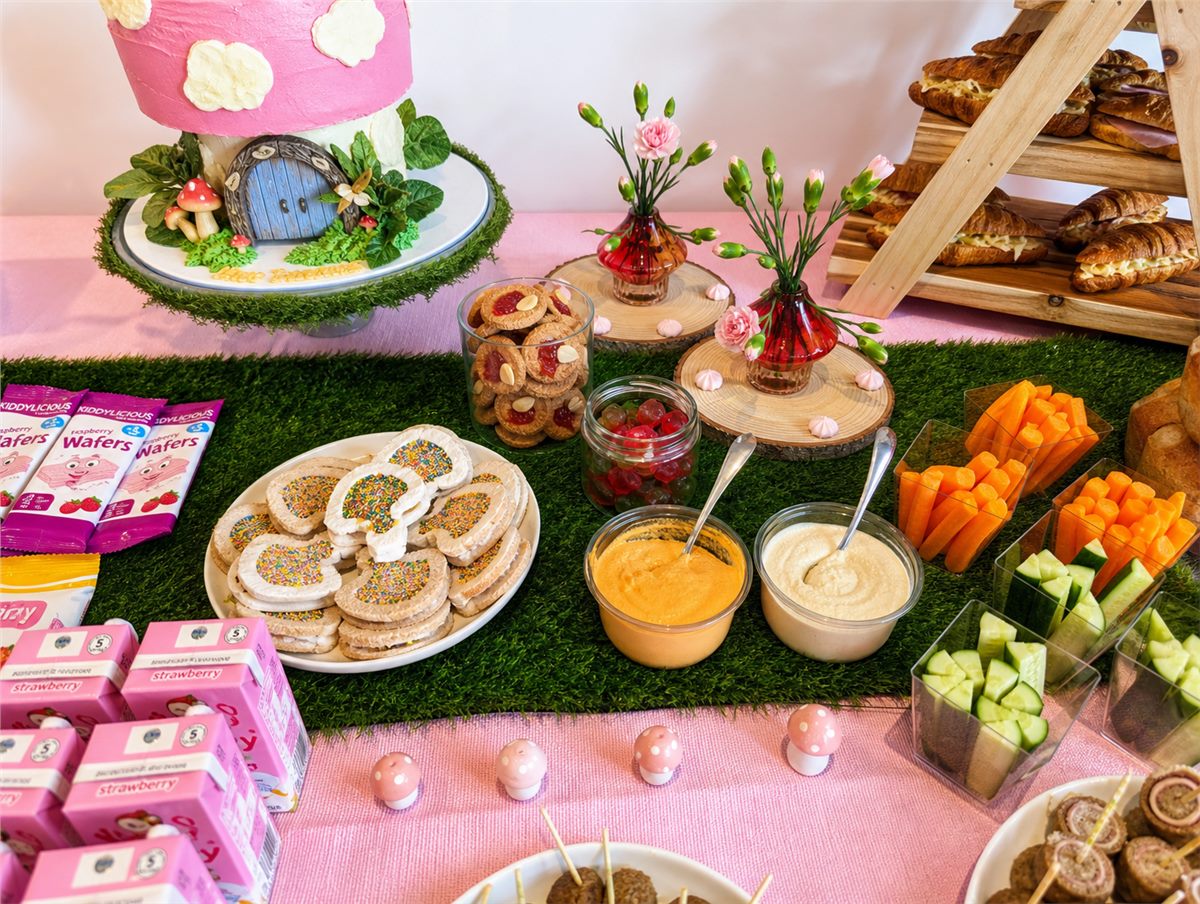 Woodland children's party food table with themed drinks, fruit, vegetables, croissants, and fairy details.